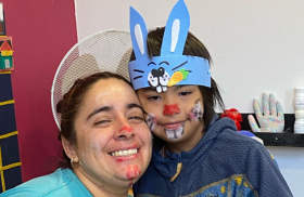 Una mujer y un niño sonríen juntos, ambos con la cara pintada como conejos. La mujer lleva una camiseta azul claro y tiene la nariz roja pintada. El niño lleva una diadema azul con orejas de conejo y la cara decorada con un bigote y nariz de conejo. Detrás de ellos, hay una pared con decoraciones coloridas.