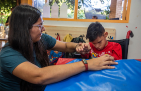 Una mujer con cabello largo y oscuro, usando gafas y camiseta verde, ayuda a un niño con una camiseta roja que está sentado en una silla de ruedas. Ambos están interactuando con una gran superficie azul acolchada. Detrás, hay una ventana que muestra árboles y luz natural.