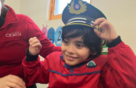 Un niño sonriente con cabello oscuro y rizado lleva una chaqueta roja y un gorro de policía con insignias doradas. Está levantando ambas manos mientras se ajusta el gorro. A su lado, una persona con chaqueta roja lo acompaña. De fondo, se ven una ventana y decoraciones coloridas con números.