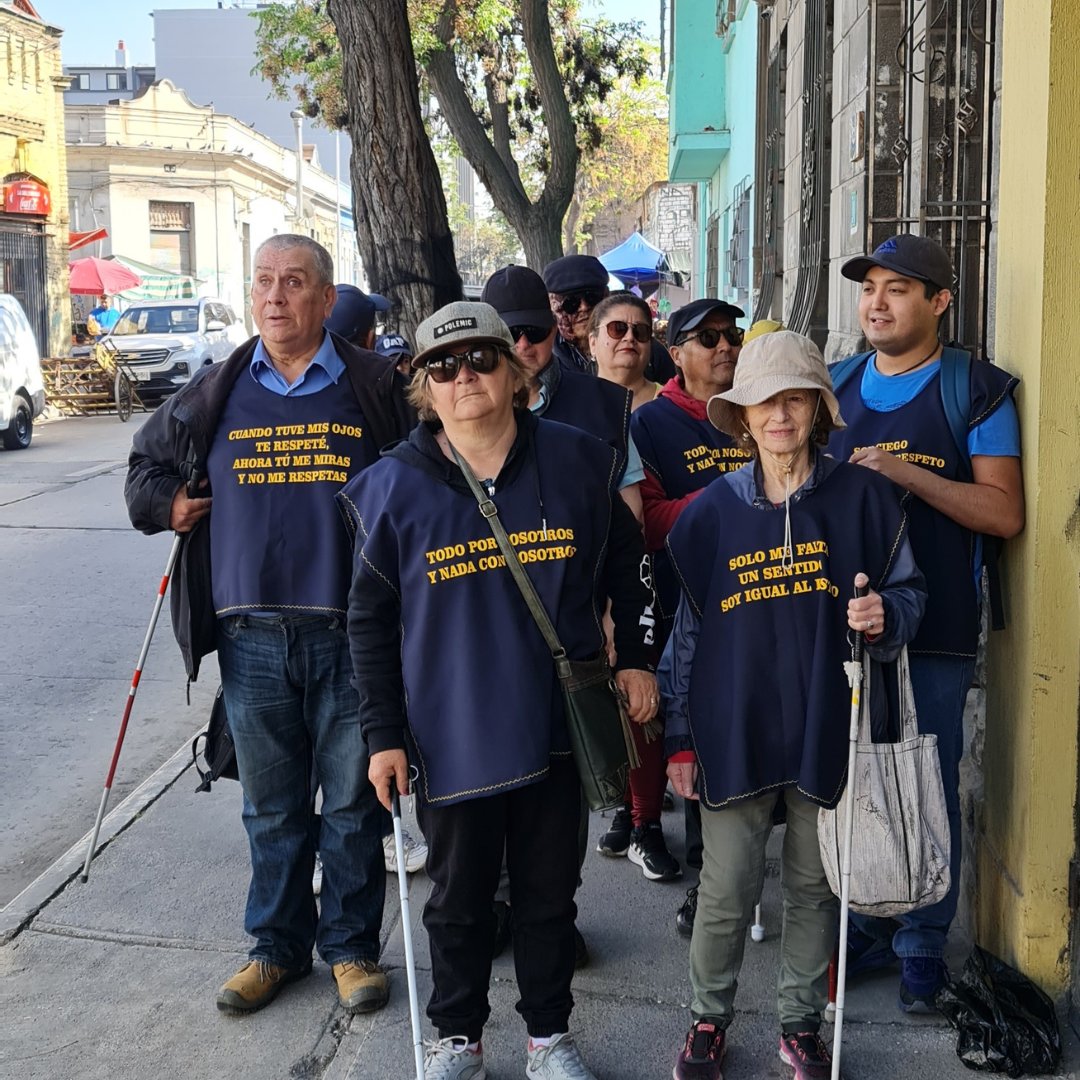 Un grupo de personas con discapacidad visual camina en fila por la calle, llevando bastones blancos y chalecos con frases que promueven respeto e inclusión hacia las personas ciegas.