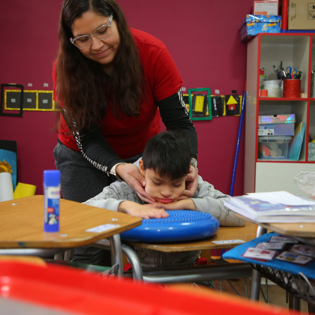 Una mujer ayuda a un niño en un aula escolar. El niño está sentado con la cabeza apoyada en sus manos sobre un cojín azul, mientras la mujer lo guía suavemente sosteniéndolo por la cabeza.