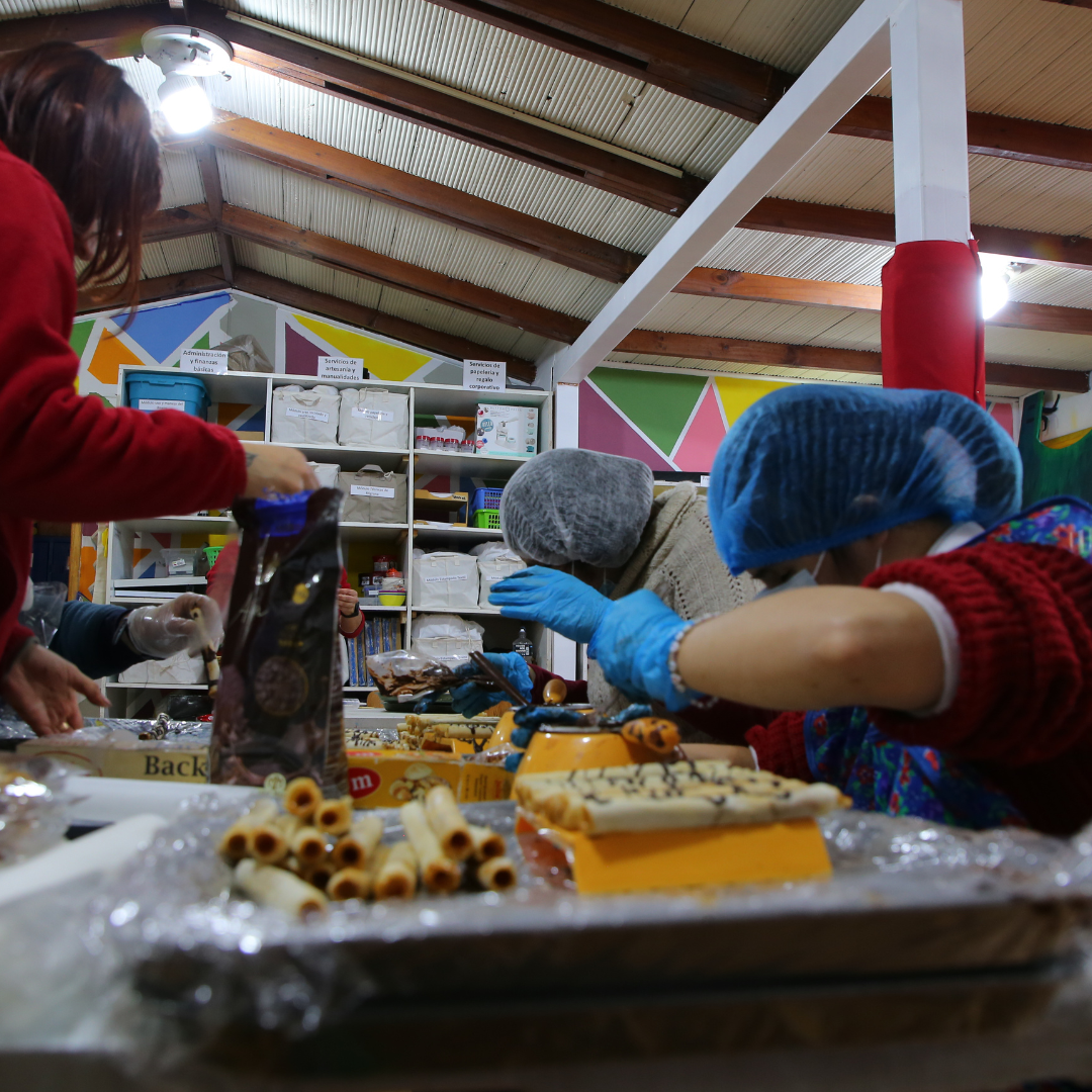 Varias personas, usando guantes y gorros de cocina, preparan alimentos en una mesa llena de ingredientes y utensilios de repostería en un ambiente colorido y organizado, enfocados en su tarea.